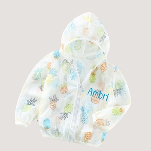 May include: White mesh jacket with a zip fastening and a hood. The jacket features a pineapple print and the name "Ambri" embroidered on the front.