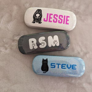 May include: Three different eyeglass cases with glitter finishes. The first case is silver with a pink glitter finish and says "JESSIE". The second case is black with a silver glitter finish and says "RSM". The third case is blue with a silver glitter finish and says "STEVE".