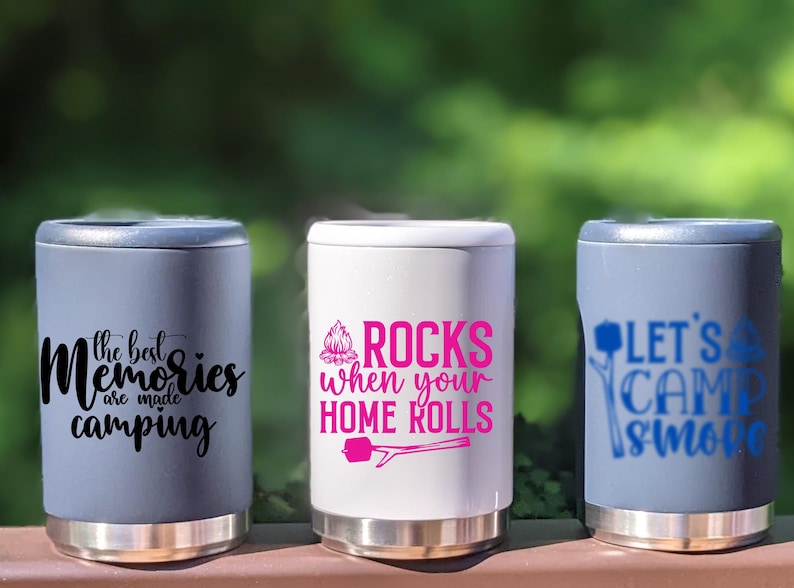 Puede incluir: Tres enfriadores de latas de acero inoxidable con diferentes dise&ntilde;os de tem&aacute;tica de campamento. El primer enfriador es gris con el texto "the best memories are made camping". El segundo enfriador es blanco con el texto "ROCKS when your HOME ROLLS" y un gr&aacute;fico de fogata rosa. El tercer enfriador es gris con el texto "LET'S CAMP SMORE" y un gr&aacute;fico de fogata azul.