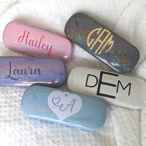 May include: Five different eyeglass cases with glitter finishes. The cases are personalised with names and initials. The names include Hailey, Laura, DEM, CAM, and a heart with the initials EA.