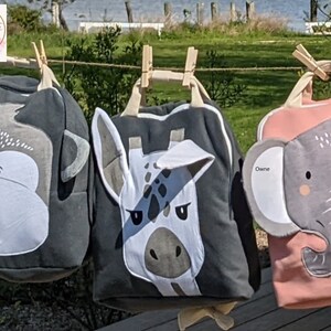 May include: Three gray and pink fabric bags with animal faces. The bags have a monkey, giraffe, and elephant face on them. The bags are hanging on a clothesline with wooden clothespins.