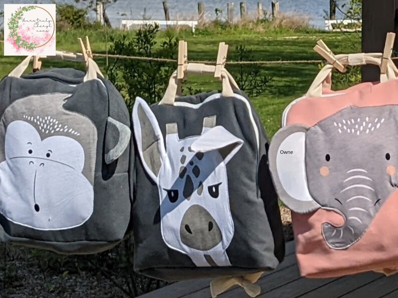 May include: Three gray and pink fabric bags with animal faces. The bags have a monkey, giraffe, and elephant face on them. The bags are hanging on a clothesline with wooden clothespins.