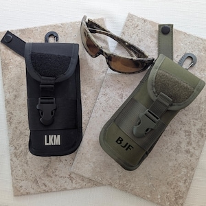 May include: Black and olive green tactical sunglass cases with personalized initials. The cases have a hook, a buckle closure, and a loop for attachment. Camouflage sunglasses rest on a neutral surface.