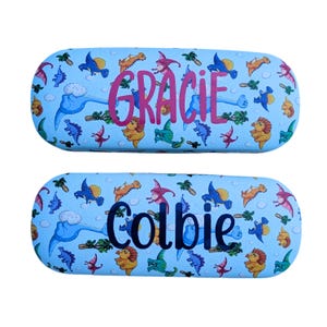 Peut inclure: Deux étuis à lunettes bleu clair avec un motif de dinosaures. L'étui supérieur porte le nom "Gracie" en rose, et l'étui inférieur porte le nom "Colbie" en bleu foncé. Les étuis sont recouverts de divers dinosaures de dessins animés.