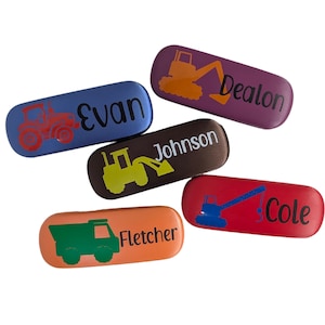 May include: Five personalized eyeglass cases with different construction vehicle designs and names. The cases are red, purple, brown, orange, and blue.