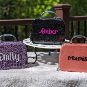 May include: Three small, textured cosmetic cases with silver handles. One is purple with "Emily" in white, another is black with "Amber" in pink, and the third is coral with "Marisa" in black. All have a reptile-like pattern.