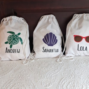 May include: Three white drawstring bags with different designs. The first bag has a green sea turtle with the name "Andrew" printed below. The second bag has a purple seashell with the name "Samantha" printed below. The third bag has red sunglasses with the name "Lola" printed below.