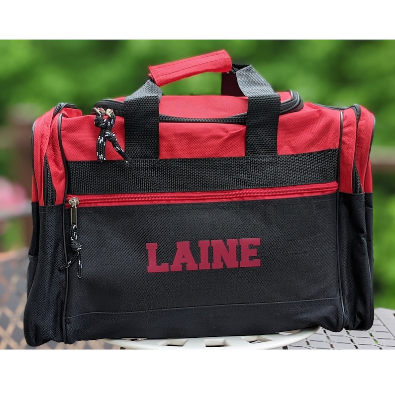 K&ouml;nnte beinhalten: Rote und schwarze Reisetasche mit Rei&szlig;verschluss und dem Namen "LAINE" in Rotbraun auf der Vorderseite.