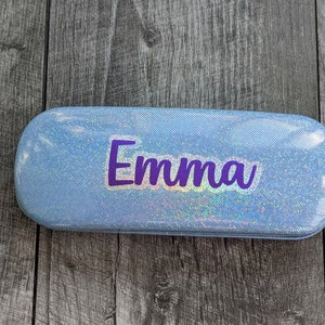 May include: A light blue, iridescent, rectangular glasses case with the name "Emma" printed in purple.