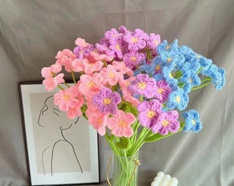 May include: A bouquet of handmade crochet flowers in a clear glass vase. The flowers are in shades of pink, purple, and blue with yellow centers. The stems are green. A framed art print and a white candle are in the background.