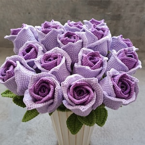 May include: A bouquet of crocheted purple roses in a white vase. The roses are made with a soft, textured yarn and have a realistic appearance.