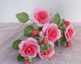 May include: A bouquet of crocheted pink and red roses with green leaves and buds. The roses are arranged in a cluster and are sitting on a white background.