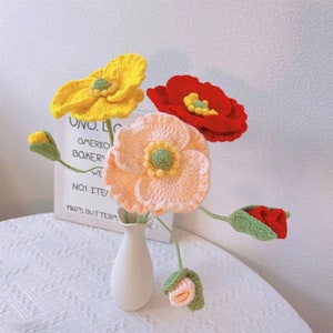 May include: A bouquet of crocheted flowers in a white vase. The flowers are yellow, pink, and red. The vase is on a white table with a white and gray patterned tablecloth.