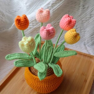 Pode incluir: Um arranjo de flores de crochê com tulipas rosa, amarelas e laranja em um vaso marrom e laranja. As flores estão dispostas em um vaso com folhas verdes.