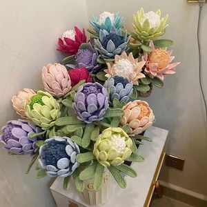 May include: A bouquet of crocheted protea flowers in a variety of colours, including pink, purple, blue, green and yellow. The flowers are arranged in a vase with a striped pattern.