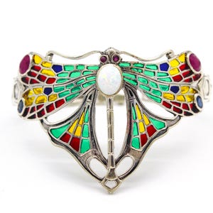 May include: A silver-toned metal cuff bracelet shaped like a dragonfly. The wings feature translucent enamel panels in green, yellow, red, and blue. The body is accented with a central oval white cabochon and two small round red stones at the head.