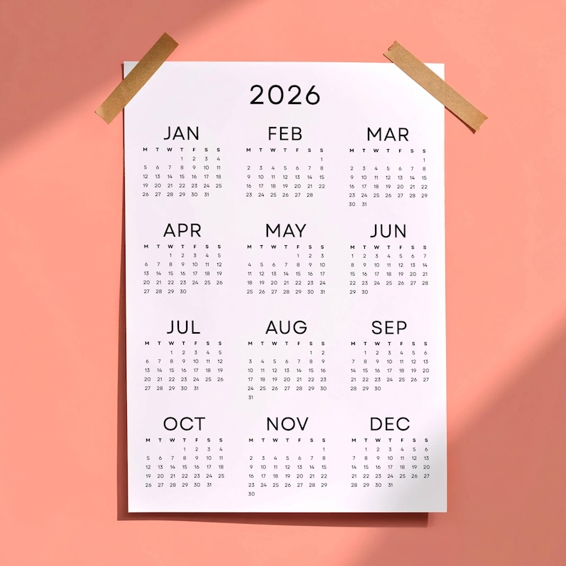May include: A white 2026 calendar with a monthly layout, taped to a pink background. Each month is displayed with its days and dates in a simple, easy-to-read format. The calendar is held in place with two pieces of tan tape.