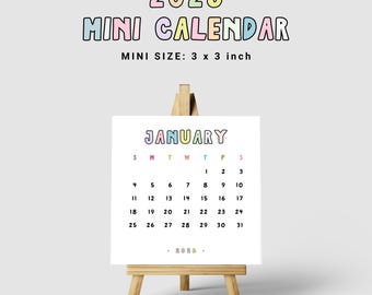 May include: A mini 2026 calendar, 3 x 3 inches, displayed on a small wooden easel. The calendar for January is visible, with colorful text and a white background. The text "2026 MINI CALENDAR" is at the top.