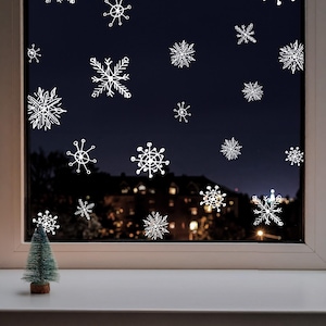 May include: A window with white snowflake decals against a dark blue background, with a small green Christmas tree on the windowsill. The window overlooks a cityscape with lights.