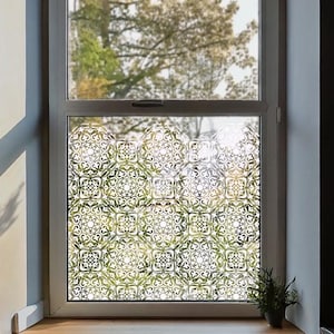 Könnte beinhalten: Ein Fenster mit dekorativer, gemusterter Folie. Die Folie zeigt ein sich wiederholendes Blumendesign in Weiß auf einem durchscheinenden Hintergrund. Das Fenster befindet sich in einem dunklen Raum mit einer Holzfensterbank und einem Heizkörper darunter.