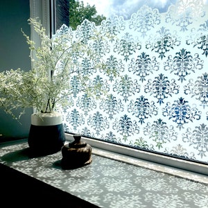 Peut inclure: Une fenêtre avec un film de confidentialité à motif damassé blanc. Le rebord de la fenêtre est décoré d'un petit objet décoratif brun foncé et d'un vase blanc et noir avec un arrangement de fleurs blanches.