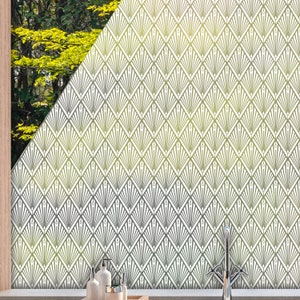 Op de afbeelding: Een witte, gematteerde glasfolie voor ramen met een geometrisch patroon van overlappende ruitvormige figuren. Het patroon is een herhalend ontwerp van gestileerde zonnestralen of waaiers. De folie is aangebracht op een raam met uitzicht op groene bomen.