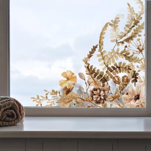 Könnte beinhalten: Ein Fensteraufkleber mit einer botanischen Illustration von Farnen, Tannenzapfen und verschiedenen Wildblumen in Braun-, Beige- und gedämpften Goldtönen. Das Design ist als Gruppe entlang der rechten Seite und der unteren Ecke der Scheibe angeordnet.