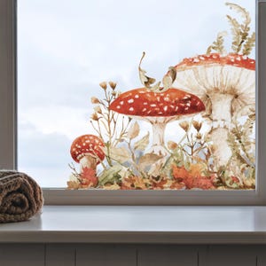 Könnte beinhalten: Ein Fensteraufkleber mit einer Aquarellillustration von drei rot-weiß gepunkteten Pilzen, umgeben von braunen Herbstblättern, getrockneten Gräsern und Farnen. Das Motiv ist auf einer Glasscheibe angebracht und zeigt erdige Farbtöne.