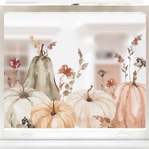 May include: A window decal featuring a watercolour illustration of four pumpkins in shades of white, cream, and muted green, accented with delicate sprigs of dried wildflowers and foliage in earthy tones of brown, rust, and soft yellow.