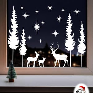 May include: A white vinyl window decal featuring a forest scene with four tall pine trees, a family of three deer, and various star shapes. The design is applied to a window pane, with a small artificial green pine tree on the sill below.