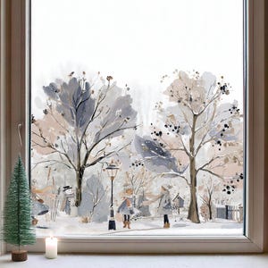 May include: A window decal featuring a winter scene with two trees, a snowman, and two figures wearing scarves and coats. The illustration uses a soft palette of grey, beige, and white tones. A small artificial green Christmas tree and a white candle sit on the windowsill in front of the glass.