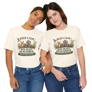 May include: Two women wearing cream-colored t-shirts with a graphic that reads "Keep Calm Wildlife Rehabber." The graphic features illustrations of various animals. The text "It's my job to save the cute & the critical!" is also included.