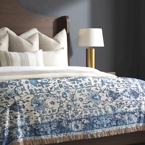 Puede incluir: Una cama con cabecero de madera oscura, cubierta con almohadas blancas y una manta con estampado floral azul y blanco con bordes de flecos. Una lámpara de mesa dorada con pantalla blanca se sitúa en una mesita de noche.
