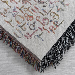 Close-up of the corner of a cream Armenian bird-letter blanket, showing colorful woven lettering and dark fringe.