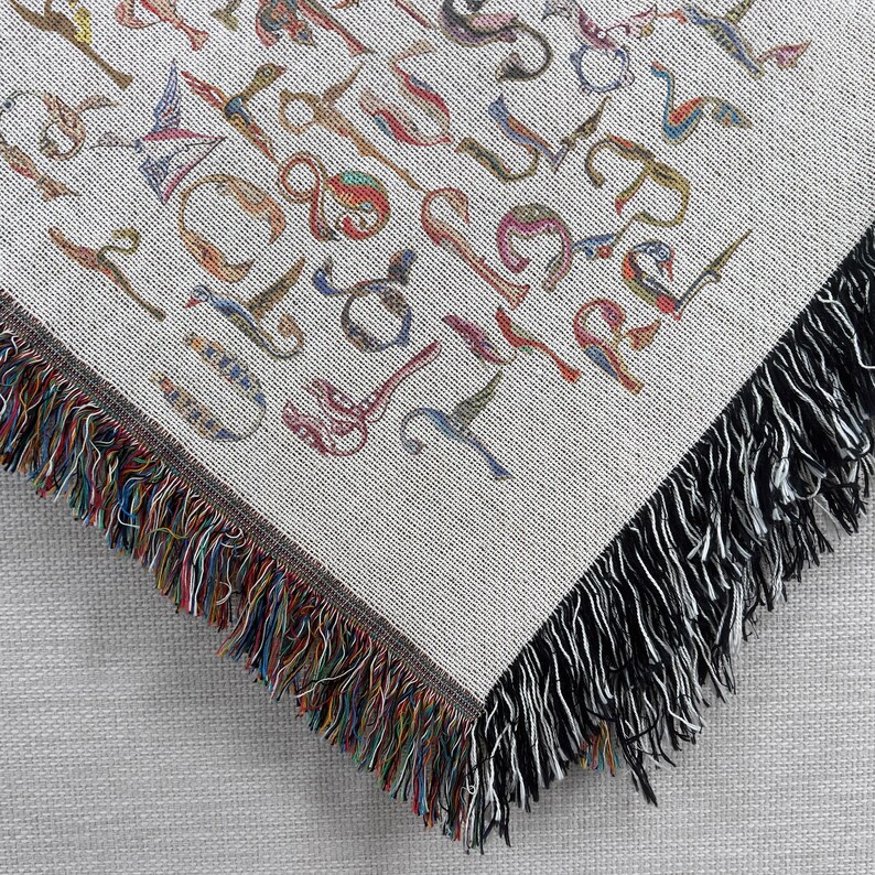 Close-up of the corner of a cream Armenian bird-letter blanket, showing colorful woven lettering and dark fringe.