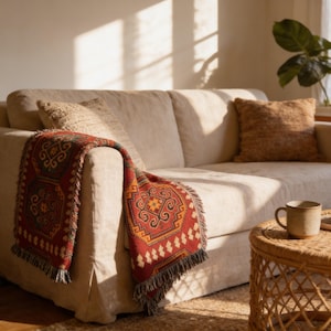 a cozy living room with a beige sofa adorned with a red and orange patterned throw blanket draped over the armrest. A woven coffee table sits in front of the sofa, and a potted plant is visible in the background.