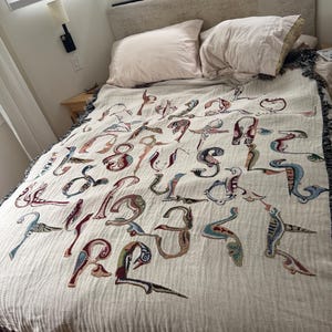 May include: A cream-colored woven throw blanket featuring a repeating pattern of stylized, colorful bird-shaped letters of the alphabet. The blanket has a textured surface and dark, fringed edges, draped over a bed with two light-colored pillows.