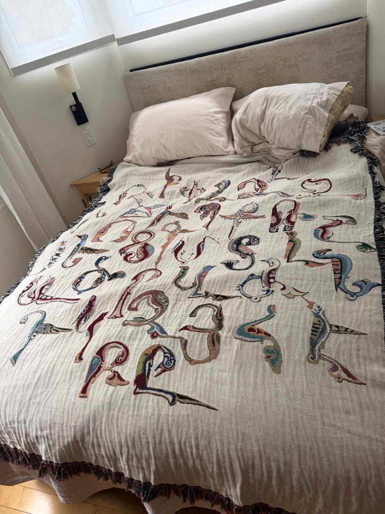 May include: A cream-colored woven throw blanket featuring a repeating pattern of stylized, colorful bird-shaped letters of the alphabet. The blanket has a textured surface and dark, fringed edges, draped over a bed with two light-colored pillows.