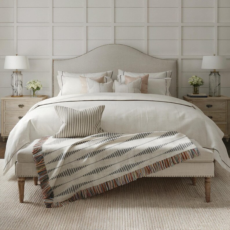 Turquoise, rust and cream patterned fringed blanket folded across the foot of a bed with neutral pillows in a bright bedroom.