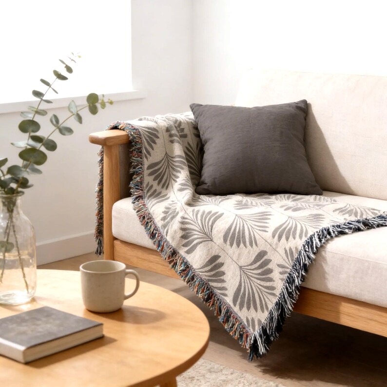 a cozy living room with a beige sofa, a wooden coffee table, and a gray throw blanket with a leaf pattern. A white mug and a book are also present on the table.