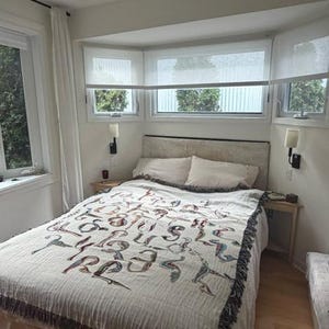 May include: A woven throw blanket featuring a repeating pattern of stylized, colorful abstract shapes and lines in shades of red, blue, and brown, draped over a bed with white pillows and a light-colored headboard in a bedroom setting.