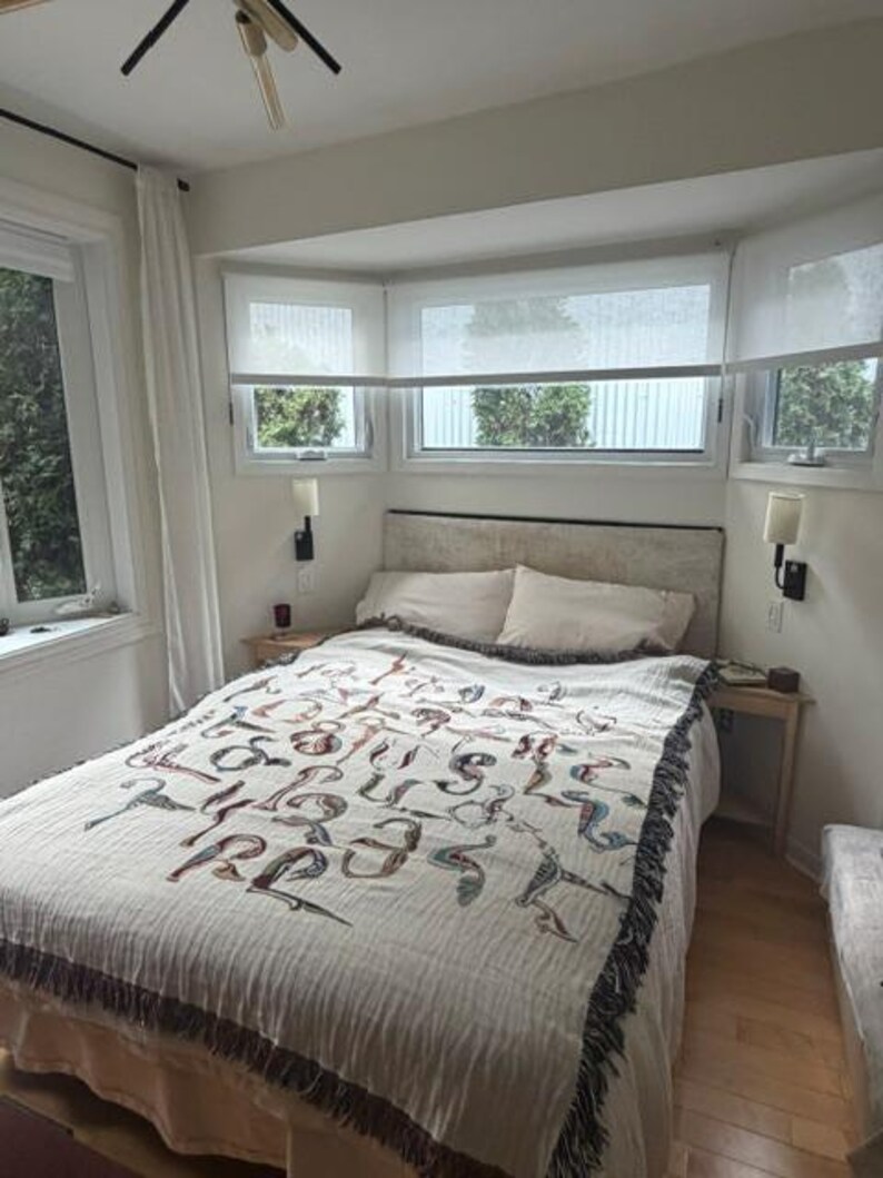 May include: A woven throw blanket featuring a repeating pattern of stylized, colorful abstract shapes and lines in shades of red, blue, and brown, draped over a bed with white pillows and a light-colored headboard in a bedroom setting.