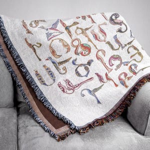 Close view of a cream Armenian bird-letter blanket draped over a gray sofa, with colorful letters and a dark fringed border.