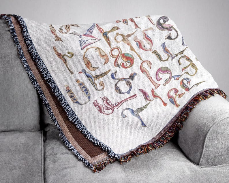 Close view of a cream Armenian bird-letter blanket draped over a gray sofa, with colorful letters and a dark fringed border.