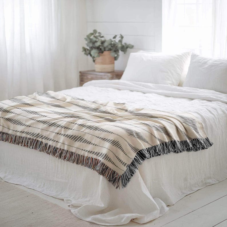 Black and cream geometric fringed throw spread across the foot of a white bed in a bright neutral bedroom.
