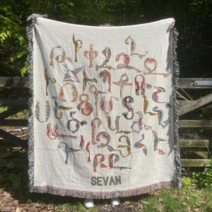 Cream woven blanket with colorful Armenian bird-shaped letters, dark fringe and a personalized name, displayed outdoors against leafy greenery.