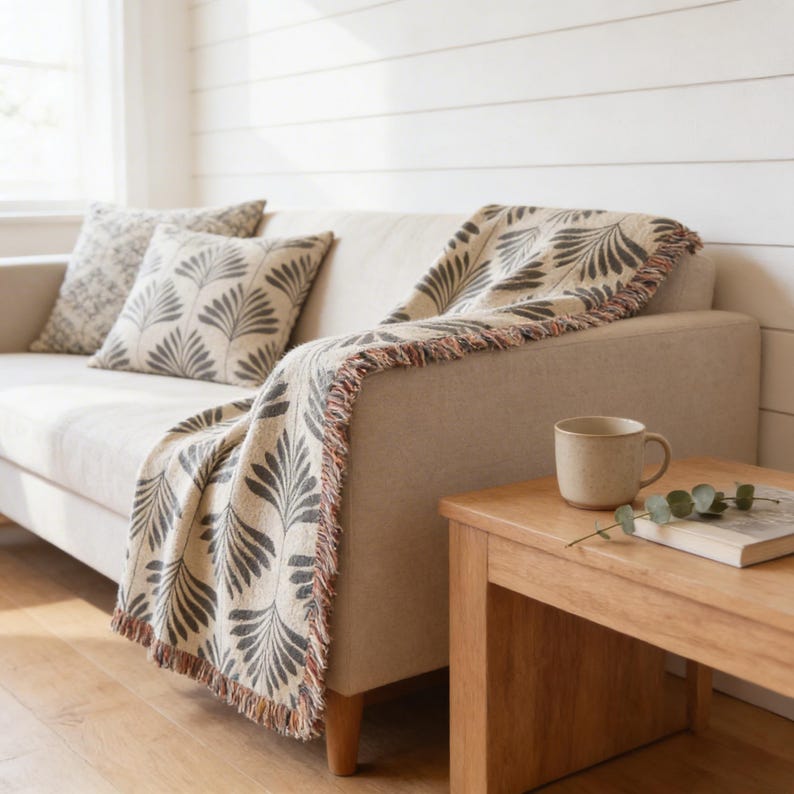 a cozy living room with a beige sofa adorned with a patterned throw blanket and two matching throw pillows. A wooden side table is positioned next to the sofa, holding a coffee mug and a book. The room has a clean, minimalist aesthetic with white walls and a wooden floor.