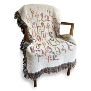 Cream Armenian bird-letter woven blanket draped over a wooden armchair, with dark fringe hanging over the seat.