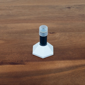 Puede incluir: Una base hexagonal blanca con un pequeño vial de vidrio negro y transparente en la parte superior.