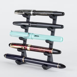 May include: A black plastic pen holder with five slots, holding five different fountain pens. The pens are black, blue, red, green, and brown.